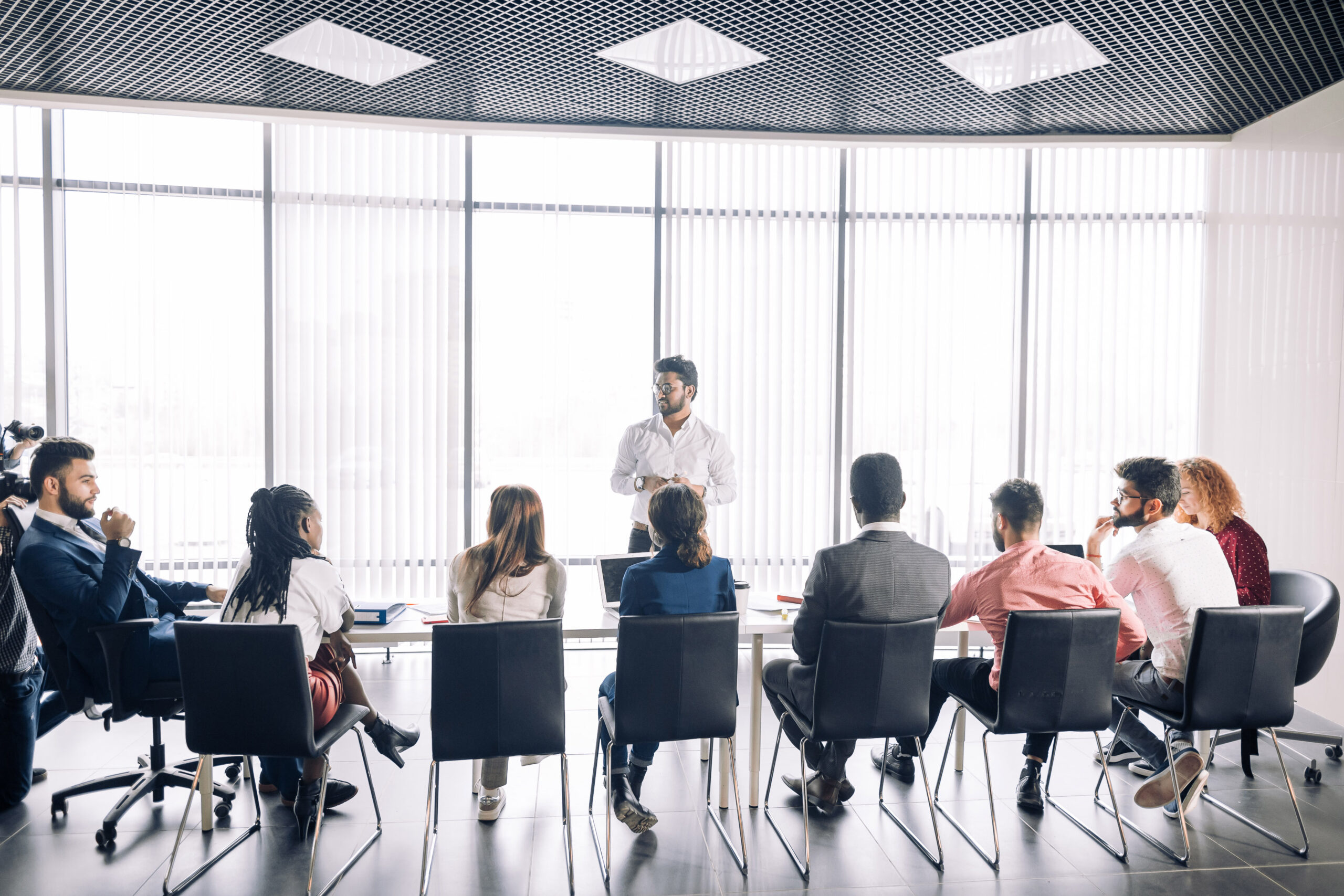 Achieving best results after annual refresher course. Indian young business mentor conducting a business training while standing in front of people sitting in row at meeting room.